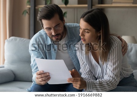 Young family couple engaged in reading loan contract mortgage agreement look through terms conditions think on signing paper. Pensive spouses husband wife embrace on sofa study legal document hardcopy Royalty-Free Stock Photo #1916669882