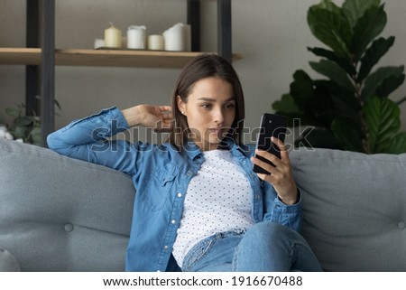 Pretty female student sit on couch hold stylish smartphone pose for cute selfie to send friend or post at social network. Pleasant young woman in trendy jeans clothes read daily news using phone app