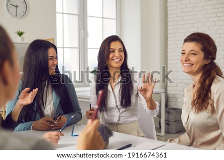 Smiling charismatic female team leader talking to employees in corporate meeting. Group of happy young business women sitting at office table, discussing ideas, sharing funny stories and laughing Royalty-Free Stock Photo #1916674361