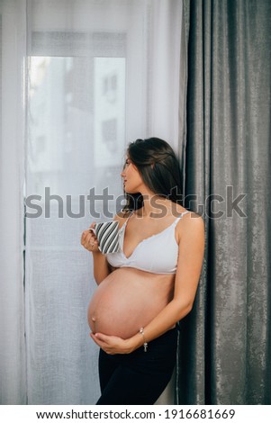 Young pregnant woman is standing by window in her apartment and holding cup of tea. 