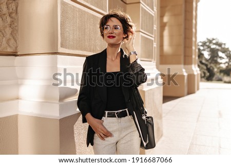 Happy girl with short hair in light trauswrs and dark jacket with handbag smiling outdoors. Modern lady in glasses posing in city.. Royalty-Free Stock Photo #1916687066