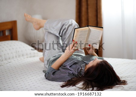 Beautiful young brunette woman in striped black and white pajamas reads a book lying on the bed in the bedroom. Royalty-Free Stock Photo #1916687132