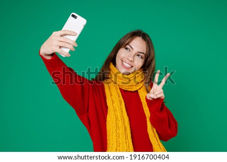 Smiling funny young brunette woman 20s wearing basic knitted red sweater yellow scarf doing selfie shot on mobile phone showing victory sign isolated on bright green color background studio portrait