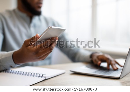 Business Application. Black Businessman Using Phone Working On Laptop At Workplace Indoors. Mobile App For Office Worker Concept. Cropped, Selective Focus On Smartphone, Shallow Depth