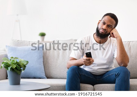 Bored African Man Watching TV Switching Television Channels With Remote Control Sitting On Couch At Home. Discontented Viewer, TV Programming Problem Concept. Free Space For Text
