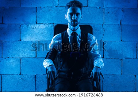 
frightened, worried, pensive, man sitting on a chair, testing on a polygraph, wearing sensors to pass a polygraph, sitting on a background of gray brickwork, looking straight. Blue filter