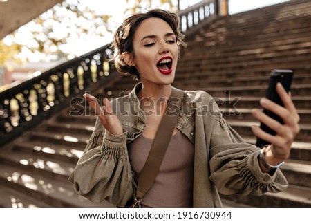 Emotional woman in long sleeve olive jacket making selfie outside. Cool girl with red lips taking photo on backdrop of stairs..
