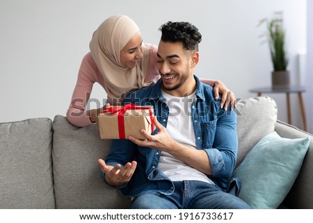 Happy muslim lady in hijab with present in her hand hugging surprised lover, home interior, free space. Emotional woman embracing her merry husband, giving Valentine gift box