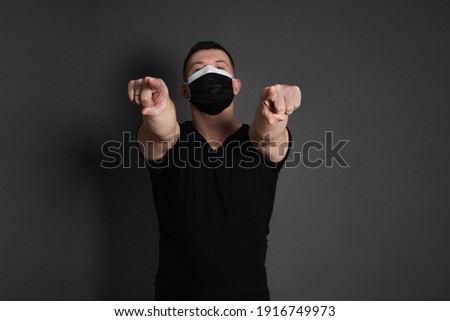 double mask. a young man, pointing with his forefingers forward, wearing a black T-shirt, against a dark background, wearing two disposable medical masks at the same time.