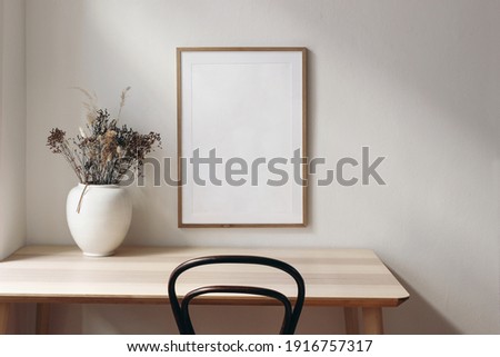 Living room, indoor still life. Empty picture frame mockup on wooden desk, table and old chair. White vase with dry grass. Elegant working space, home office concept. Scandinavian interior design.