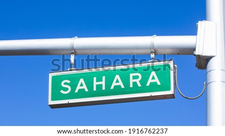 Street name sign of Sahara Avenue hanging on traffic light pole. Intersection of S Las Vegas Blvd (the Strip) where the city splits between East and West. Plain blue sky in the background.