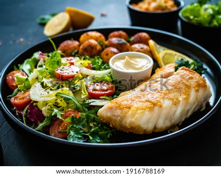 Fish dish - fried cod fillet with potatoes and vegetable salad on black wooden table