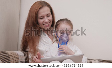 Treat your child for colds at home. Mom treats her daughter with inhalation and together they watch cartoons on tablet. Child is receiving respiratory therapy with nebulizer. Child suffers from cough.