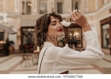 Lovely woman in white shirt with long sleeve holding flower in city. Short-haired girl with red lipstick posing at street.. Royalty-Free Stock Photo #1916867186