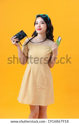Image of happy asian young woman tourist standing isolated over yellow background holding camera and passport with tickets.
