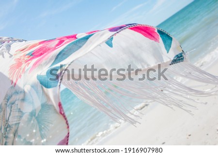 close up of a white scarf with pink and green floral pattern white fringe at a white sand beach background