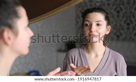 A young caucasian woman applies a cream to the skin in front of the mirror. Morning skin care treatments. Awakening from sleep concept. High quality photo