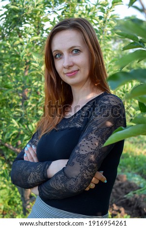 Pretty woman in black lacy blouse surrounded by apple trees stands with her hands crossed on chest. Confident beautiful girl with crossed hands poses for a photo in the apple tree garden. 
