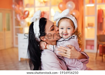 A young mother and her daughter in pajamas are laughing and having fun together in the bedroom and dressing room. A woman kisses her child.