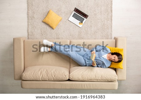Top view of young restful brunette female in blue pajamas reading book while lying on couch with head on yellow pillow after working day Royalty-Free Stock Photo #1916964383