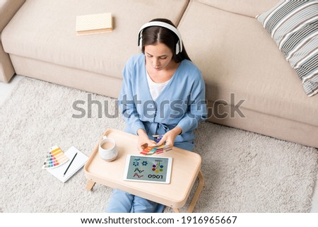 Young female designer in blue pajamas and headphones sitting on the floor by couch, listening to music and choosing colors for new logo