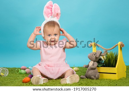 Little baby girl on green grass and blue background in the ears of an easter bunny with painted easter eggs preparing for easter