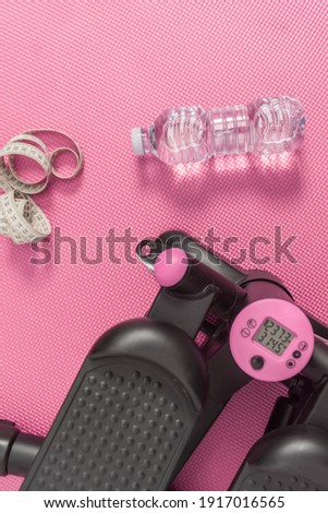Vertical photography, objects of fitness life, stepper machine to burn calories, measuring tape and water bottle on a pink background floor.