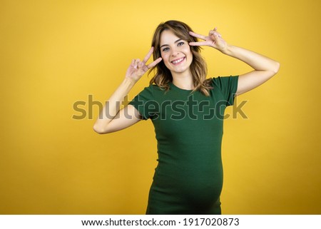 Young beautiful brunette woman pregnant expecting baby over isolated yellow background Doing peace symbol with fingers over face, smiling cheerful showing victory