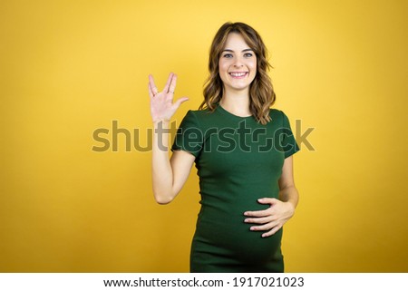 Young beautiful brunette woman pregnant expecting baby over isolated yellow background doing hand symbol