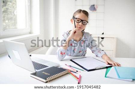 Pretty teenager girl doing homework sitting behind the table at home. Young beautiful girl with glasses, learning online with a laptop, distance learning, self-education