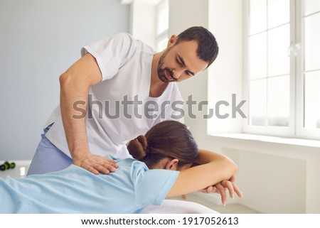 Smiling doctor osteopath in medical uniform fixing woman patients shoulder and back joints in manual therapy clinic during visit. Professional osteopath during work with patient concept Royalty-Free Stock Photo #1917052613