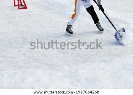 Hockey player on ice-skating removing snow with a blue shovel in the winter in a park. Wearing white clothes. Moving from goal to corner. Ice rink cleaning. Active sport outdoor