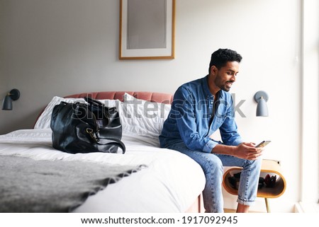 Male guest in boutique hotel sitting on edge of bed checking emails and social media on mobile phone in the morning Royalty-Free Stock Photo #1917092945