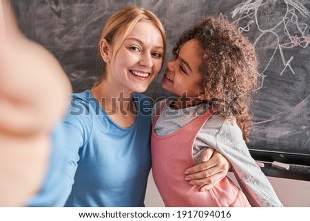 Let's make a selfie. Portrait view of the active blonde caucasian woman holding a smartphone while making a selfie with her multiracial daughter. Cute girl kissing her mother at the chick