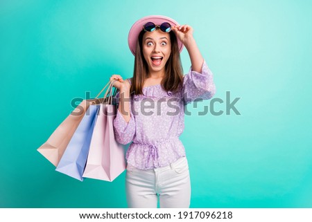 Photo of impressed brunette girl hold bags take off spectacles wear purple shirt cap trousers isolated on blue color background