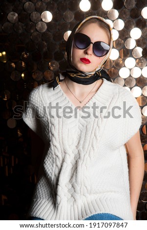 A girl in a white knitted vest, dark glasses and a silk scarf against the background of a golden wall. Fashion trends, photo shoot in the studio.
