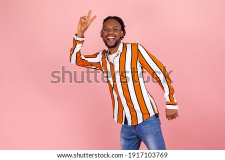 Friendly smiling african man with dreadlocks in trendy striped shirt and denim pants showing victory gesture with fingers, looking at camera. Indoor studio shot isolated on pink background