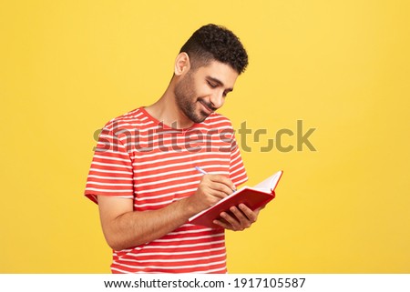 Smiling happy businessman with beard in striped t-shirt writing down to do list, making notes with pen in notebook, checking schedule. Indoor studio shot isolated on yellow background