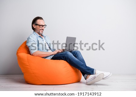 Profile side view of handsome clever cheerful mature man sitting on bean chair using laptop isolated over grey pastel color background