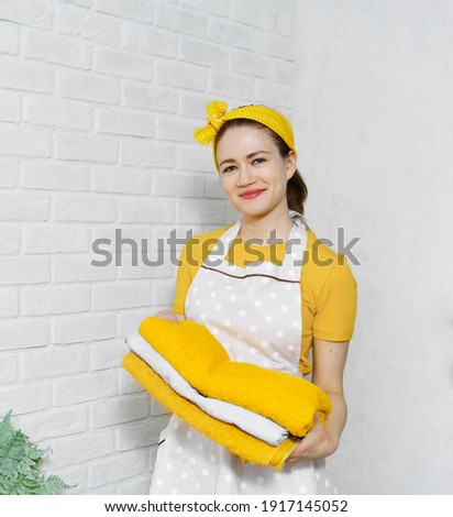 Caucasian young woman doing her homework. Housewife dressed in a yellow headband, yellow t-shirt and apron, holding the underwear on the background of a white brick wall
