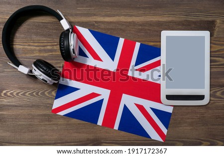 Electronic headphones and a white tablet with a blank screen lie on the flag of the United Kingdom. Close-up, brown wooden background, top view. English course online . Moskup for advertising.