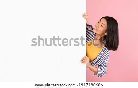 Intresting Offer. Happy Casual Asian Woman Peeping Out The Side Of White Advertisement Board And Holding It, Smiling Lady Looking At Copy Space For Your Text Or Design, Standing Over Pink Background Royalty-Free Stock Photo #1917180686
