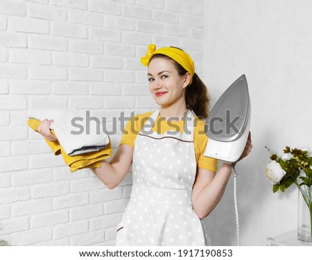 A happy young Caucasian woman in a yellow headband, yellow T-shirt and apron holding an iron and a stack of colorful underwear in her hand. Housewife washes and irons clothes