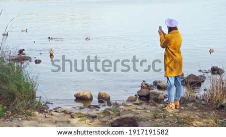 woman in trendy autumn clothes taking photo of wild ducks on lake in park