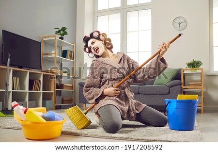 Cheerful young woman with musical talent having fun while cleaning her house. Funny crazy housewife in hair curlers and face mask tidying up home, singing loud songs and playing on pretend mop guitar