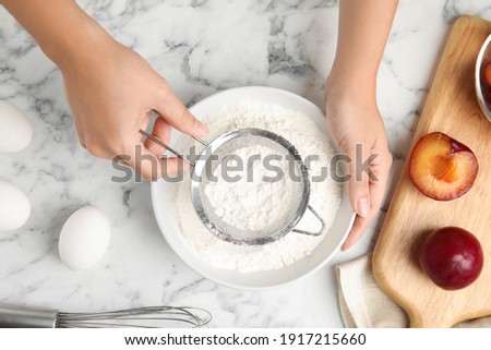 Woman sieving flour at white marble table, top view. Cooking of delicious plum cake
