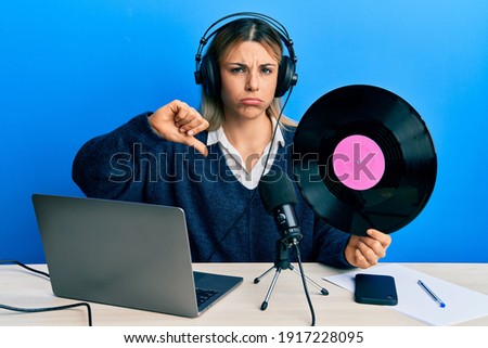 Young caucasian woman working at radio studio holding vinyl disc with angry face, negative sign showing dislike with thumbs down, rejection concept 
