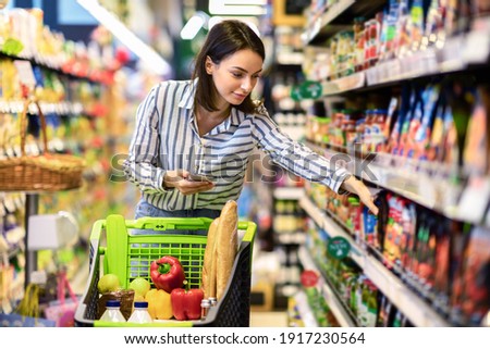 Portrait Of Millennial Lady Holding And Using Smartphone Buying Food Groceries Walking In Supermarket With Trolley Cart. Female Customer Shopping With Checklist, Taking Products From Shelf At The Shop Royalty-Free Stock Photo #1917230564
