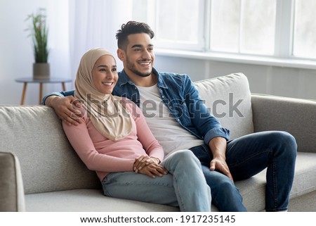 Cheerful muslim couple sitting on couch at home and looking at copy space. Handsome arab man and his wife in hijab hugging while sitting on sofa in living room, enjoying weekend together