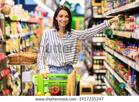 At The Supermarket. Smiling Young Woman Standing With Trolley Cart Between Aisles In Grocery Store. Cheerful Consumer Buying Essentials In Super Market, Taking Products From Shelf Looking At Camera Royalty-Free Stock Photo #1917235247